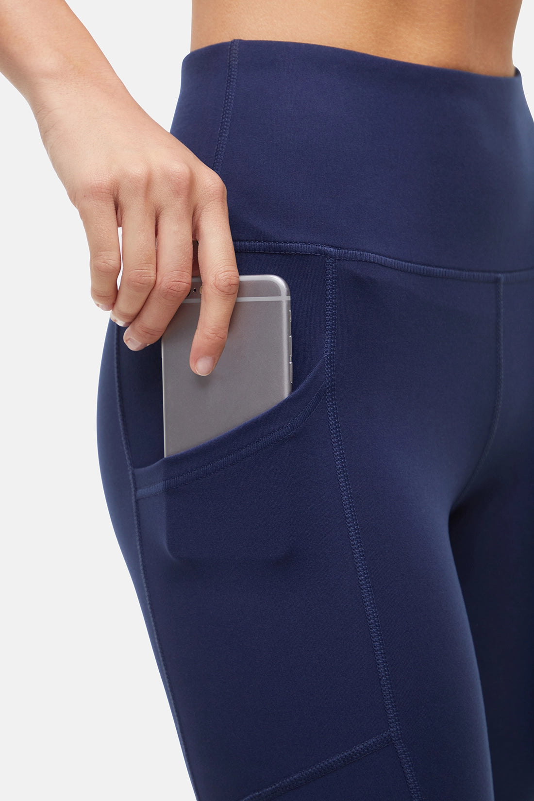 Close up side view of woman wearing navy leggings with a side pocket holding a phone on a light gray background.