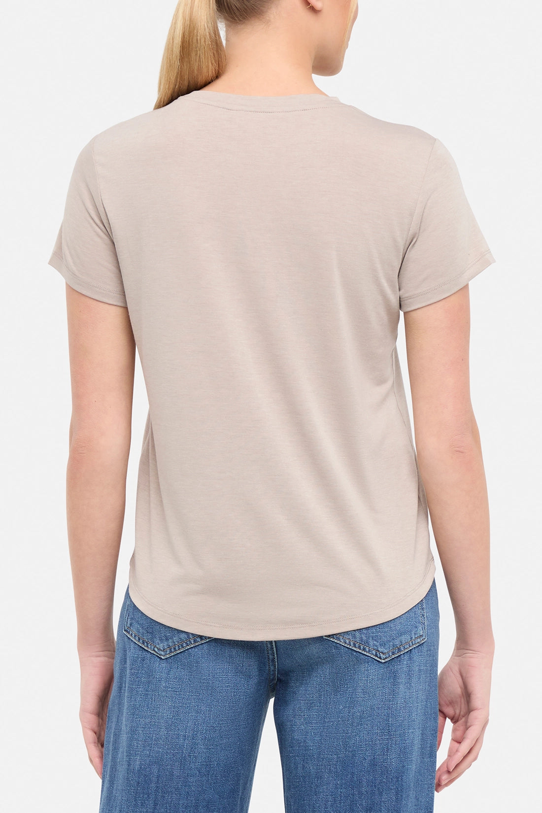 Back view of woman wearing a beige t-shirt paired with blue jeans on white background.