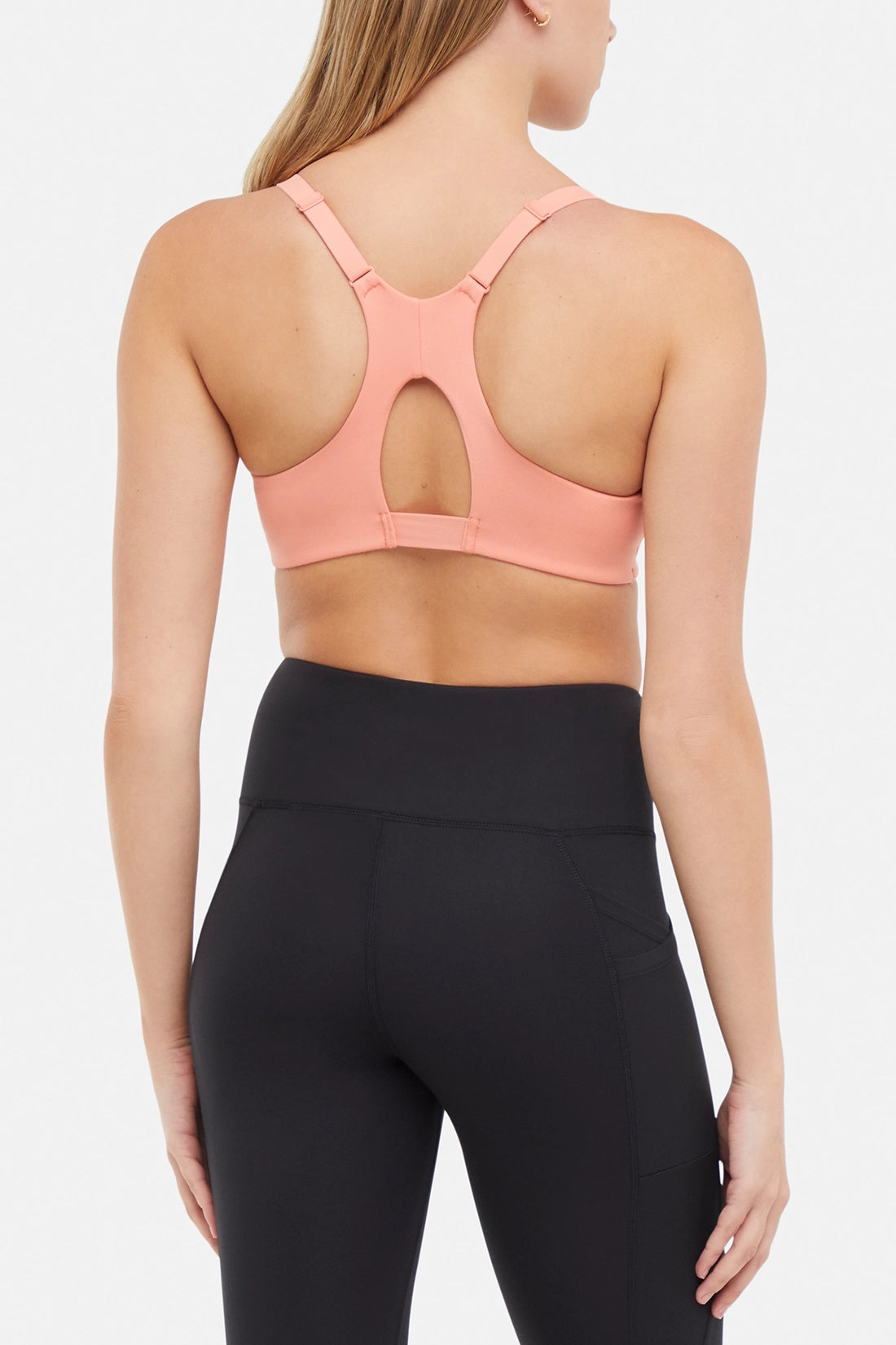 Back view of woman wearing a peach sports bra and black leggings on a white background.