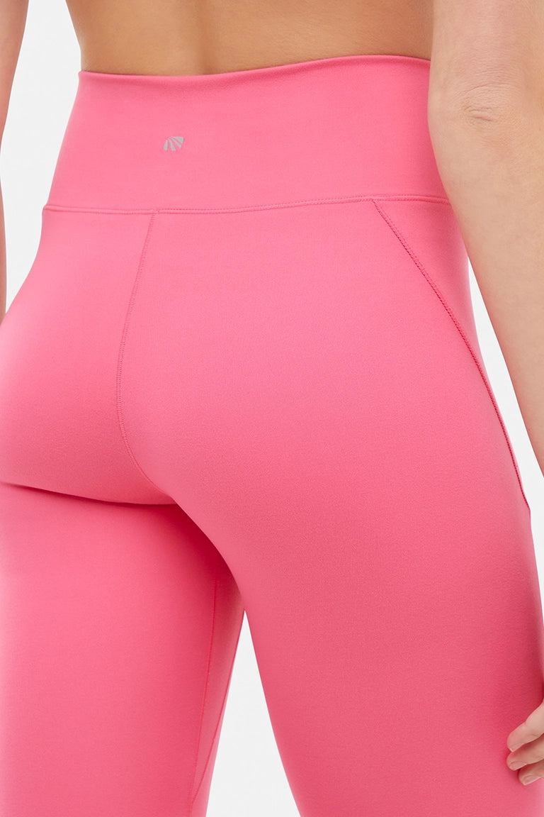 Close up back view of woman wearing pink capris with small brand logo and white sneakers on a light gray background.