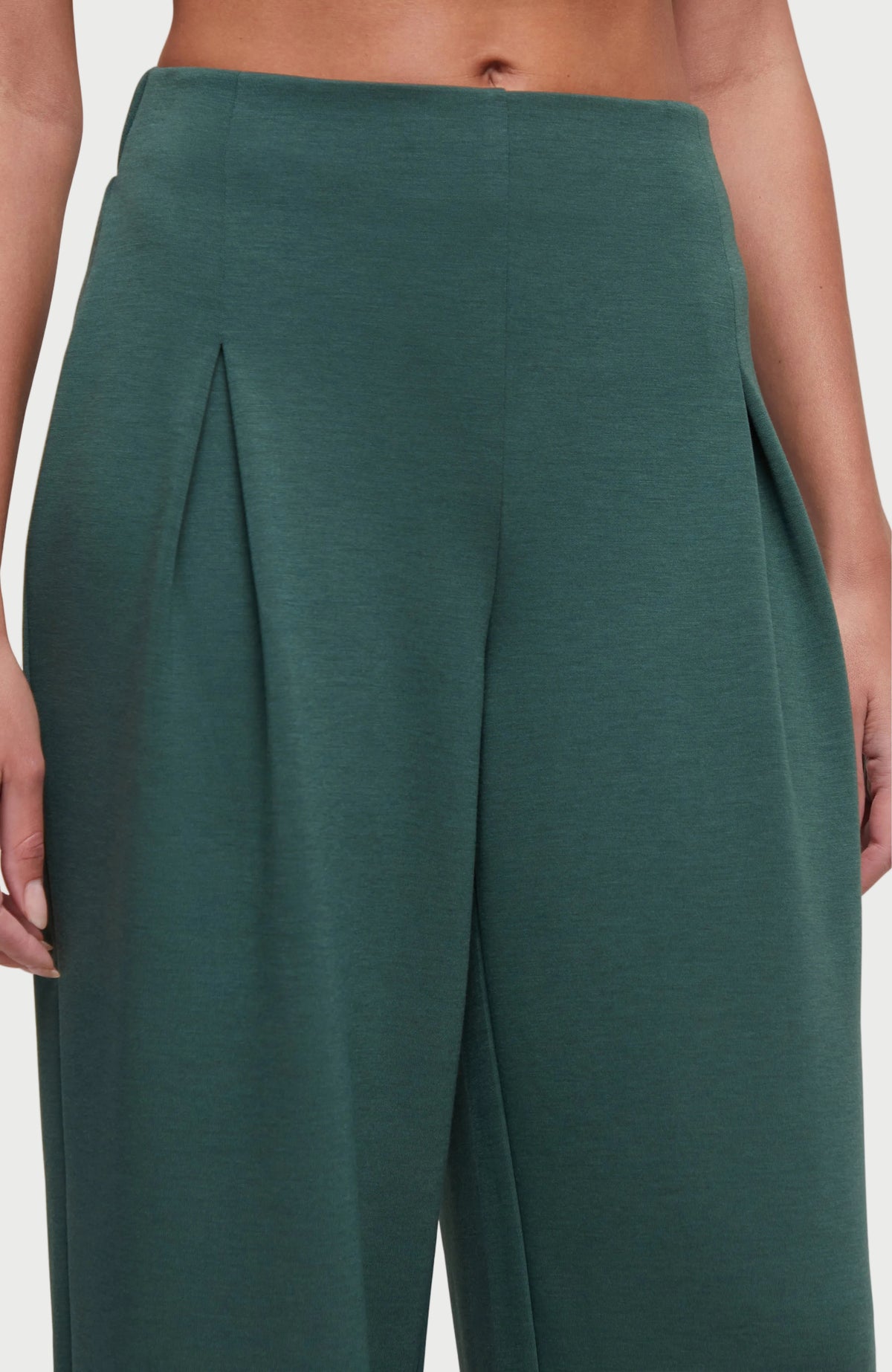 Close-up of a model wearing high-waisted, front pleats, wide-leg trousers in a deep teal green color, showing the pleated front detail and fabric texture.
