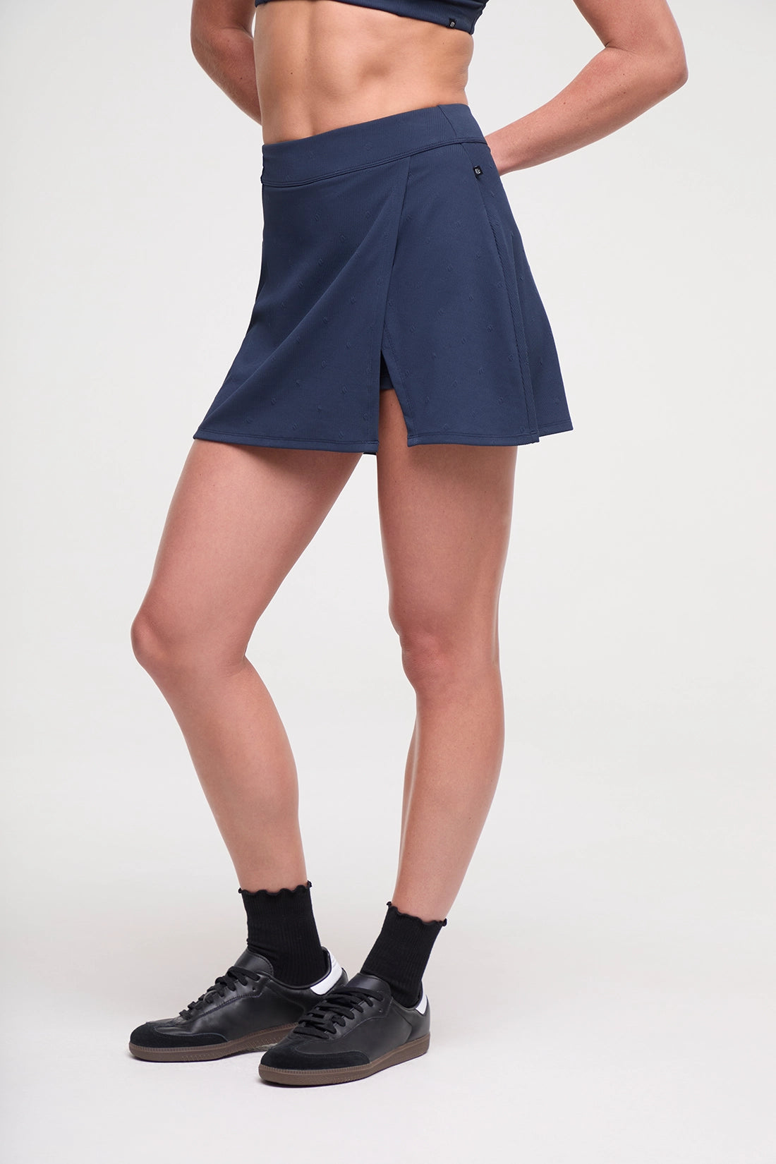 Side view of woman wearing a navy blue tennis skirt and black sneakers on a light gray background.