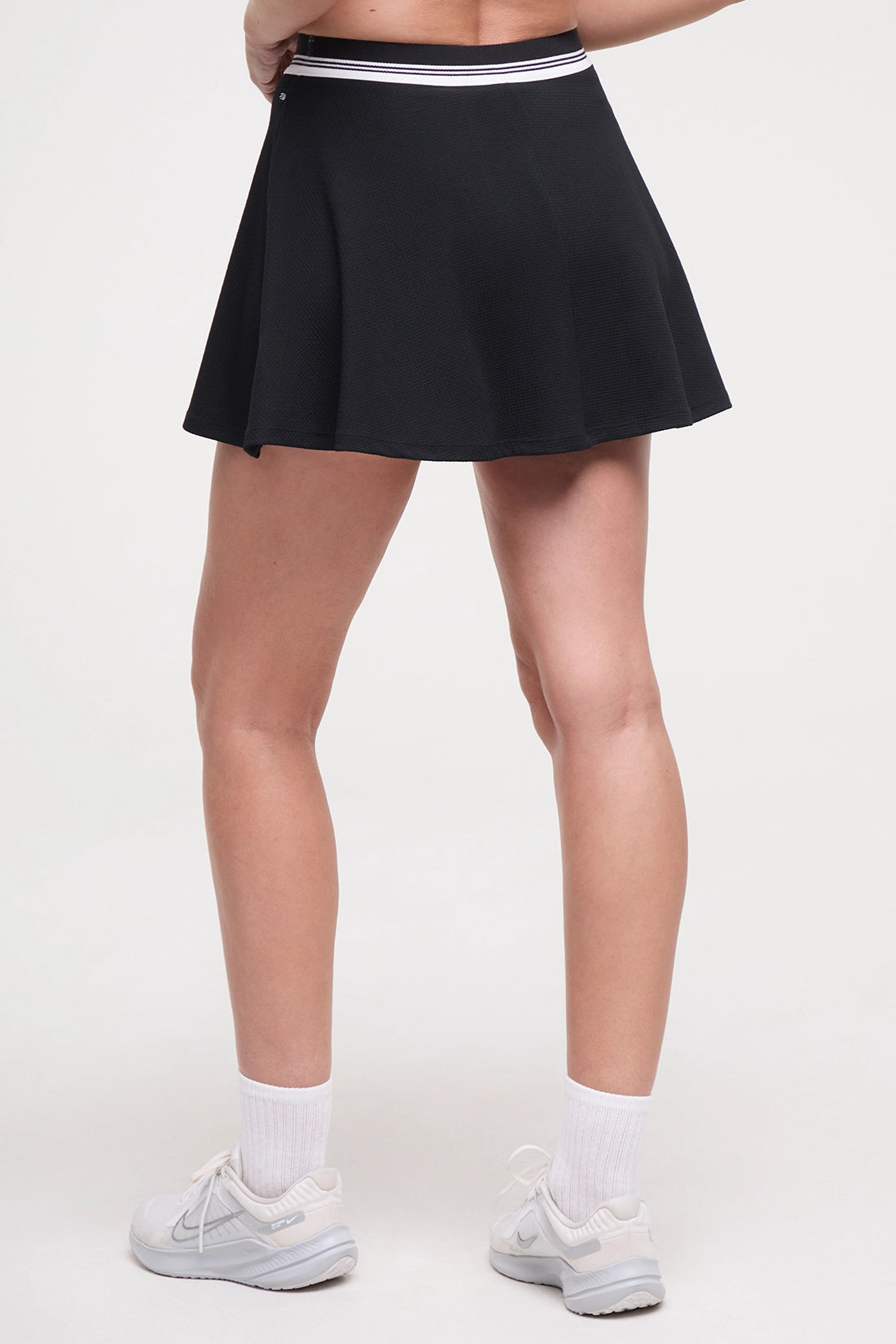 Back view of black tennis skirt with white stripe waistband worn by a woman on a white background.