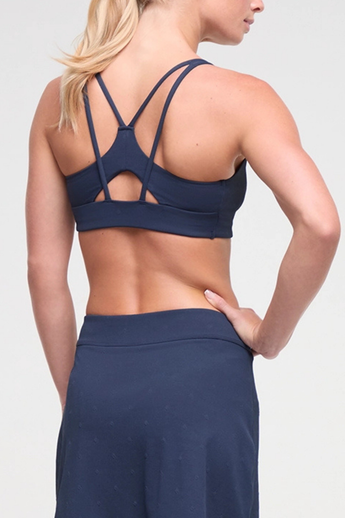 Back view of woman wearing a navy blue sports bra and skirt on a light gray background.