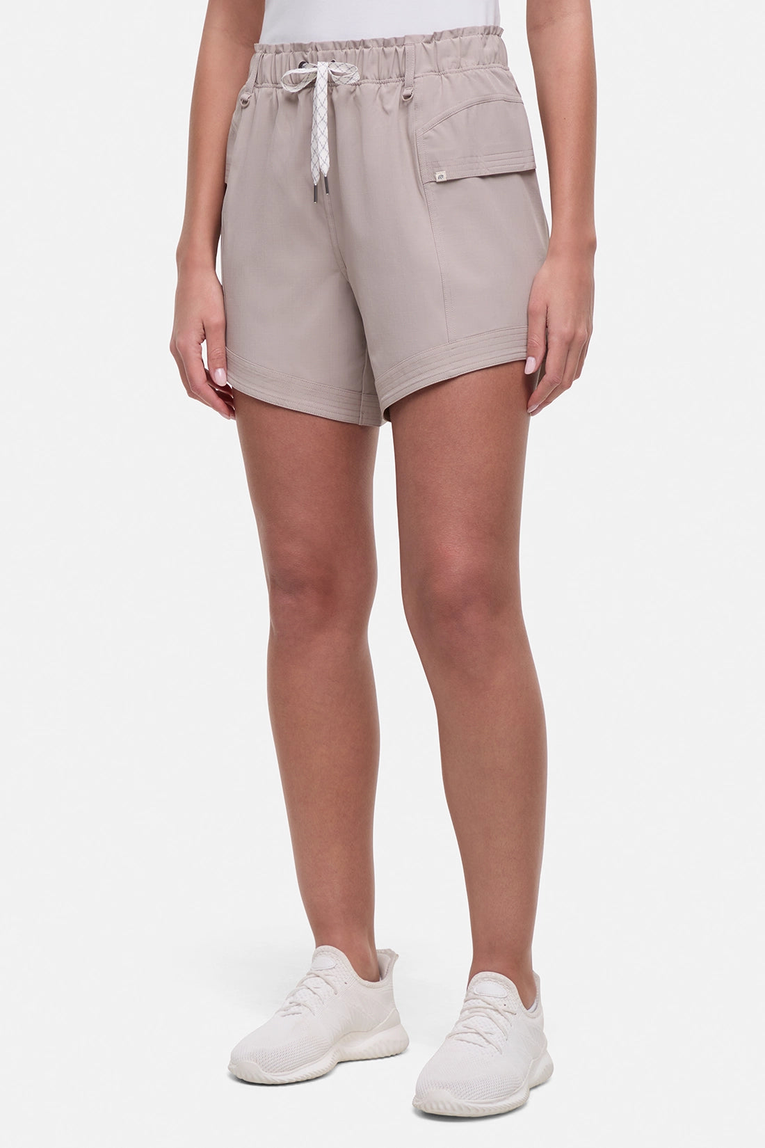 Front view of woman wearing beige shorts with white drawstring waist and side pocket and white sneakers on a white background.
