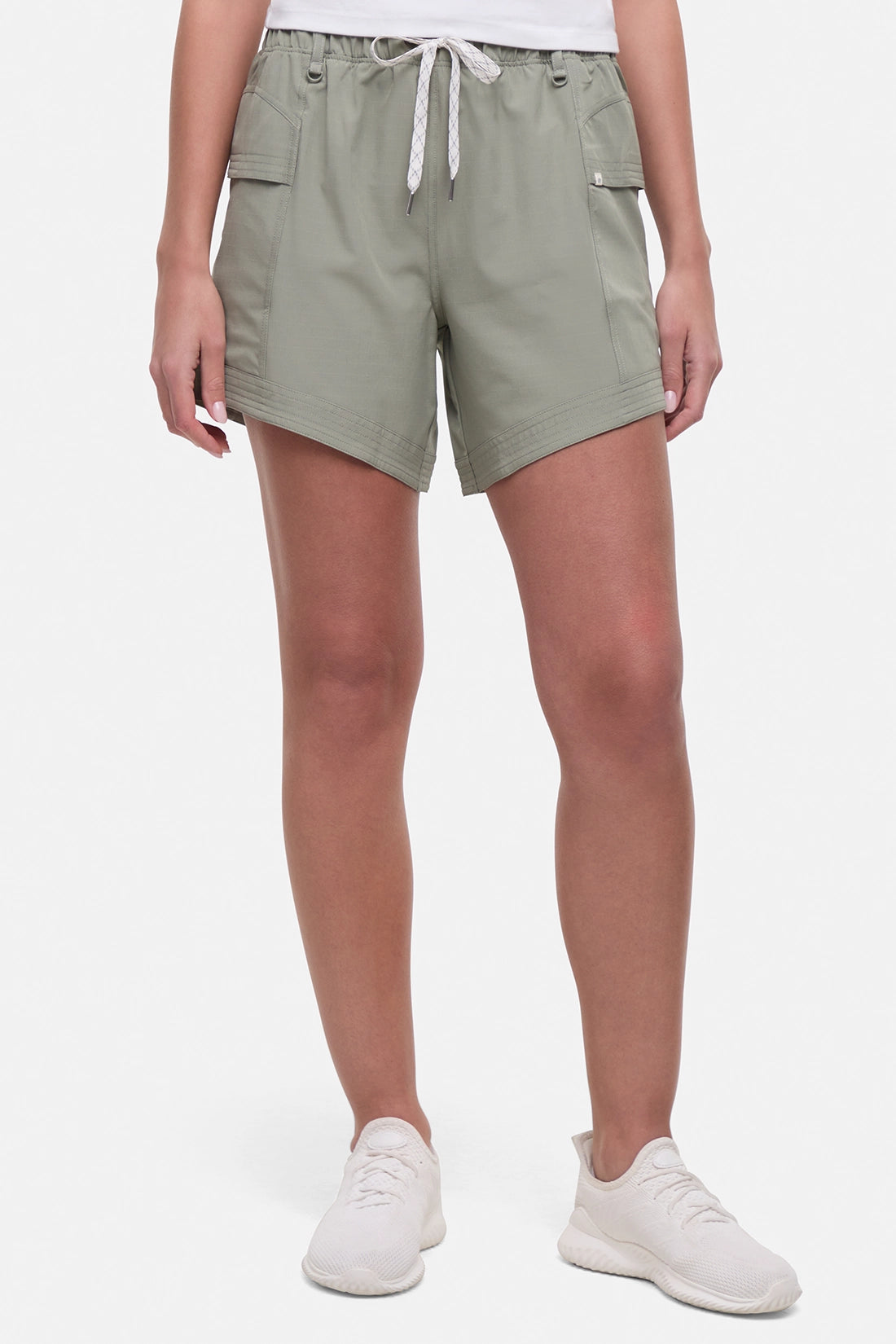 Front view of woman wearing sage green shorts with white drawstring waist and white sneakers on a white background.