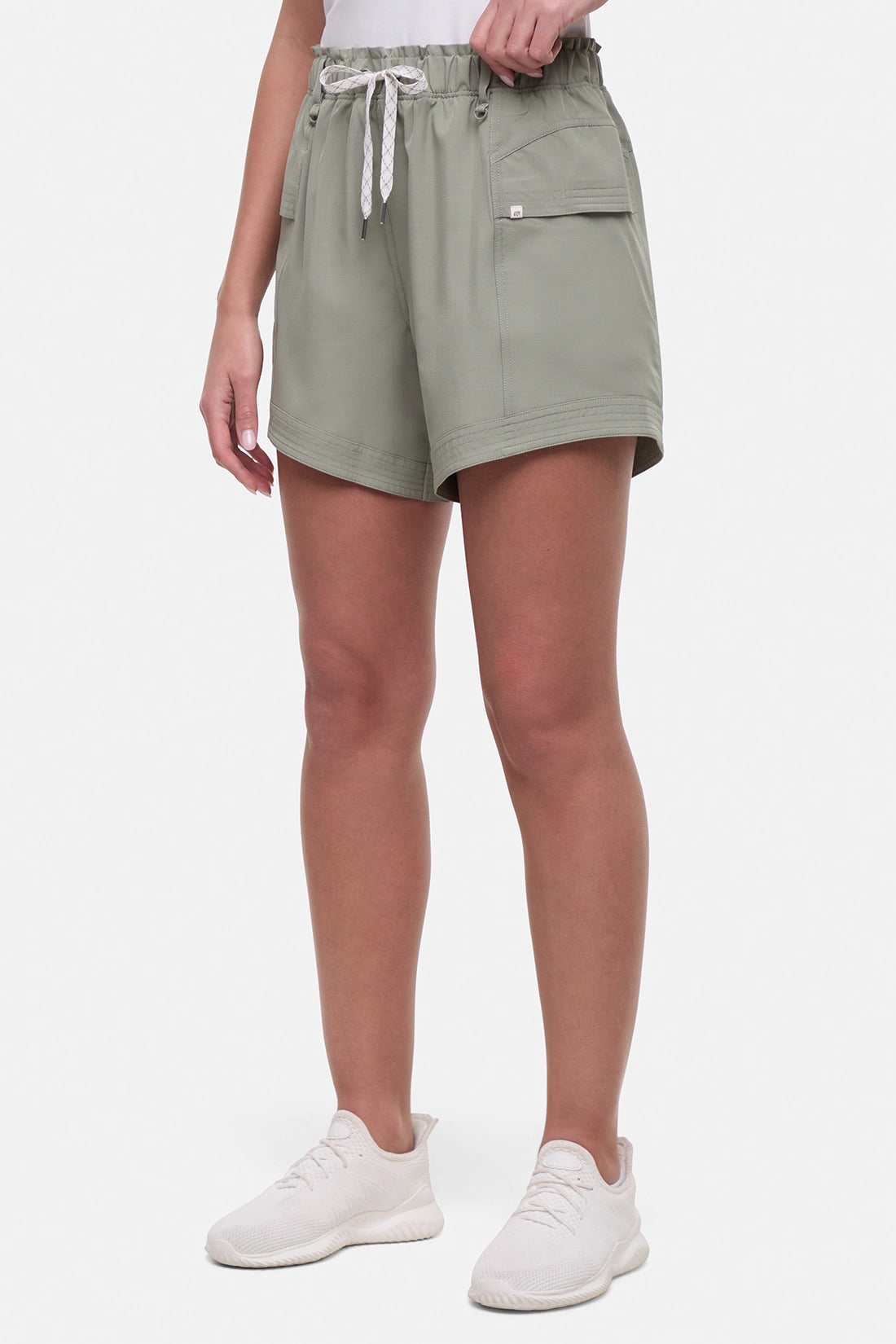 Side view of woman wearing sage green shorts with white drawstring waist and with side pockets and white sneakers on a white background.