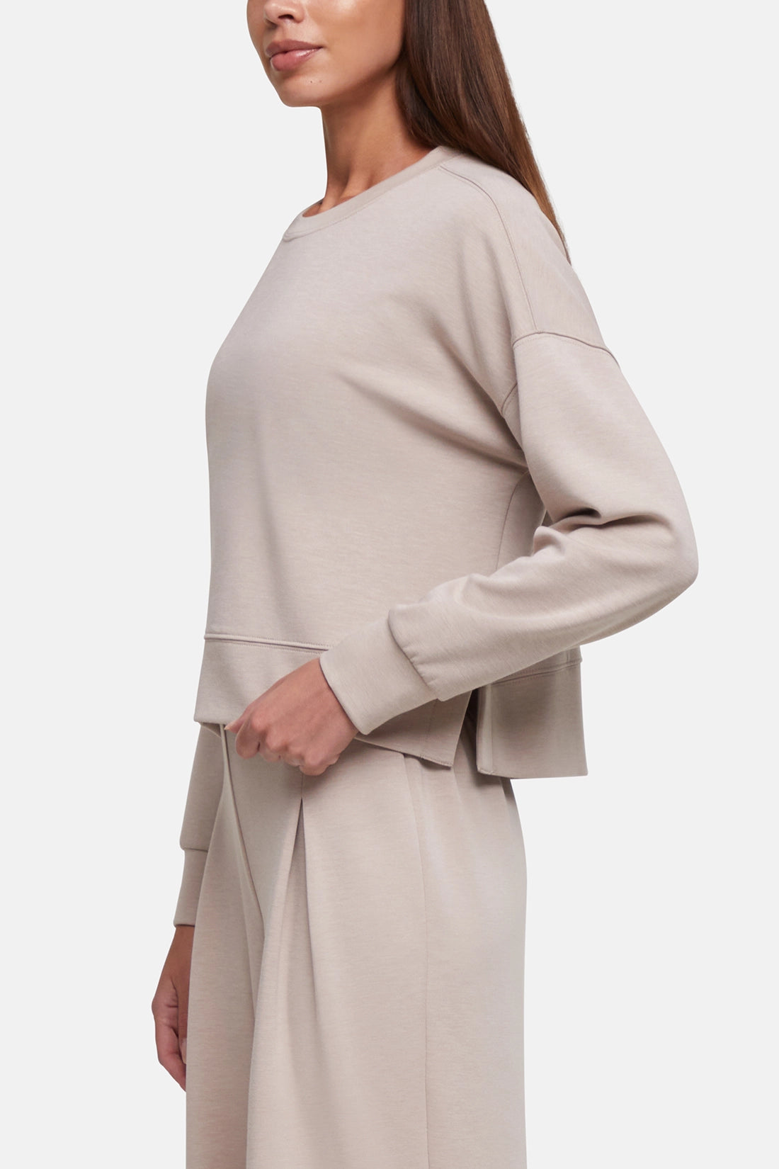 A woman wearing a light beige long-sleeve cropped sweatshirt and matching pleated skirt, shown in a side profile against a clean white background.