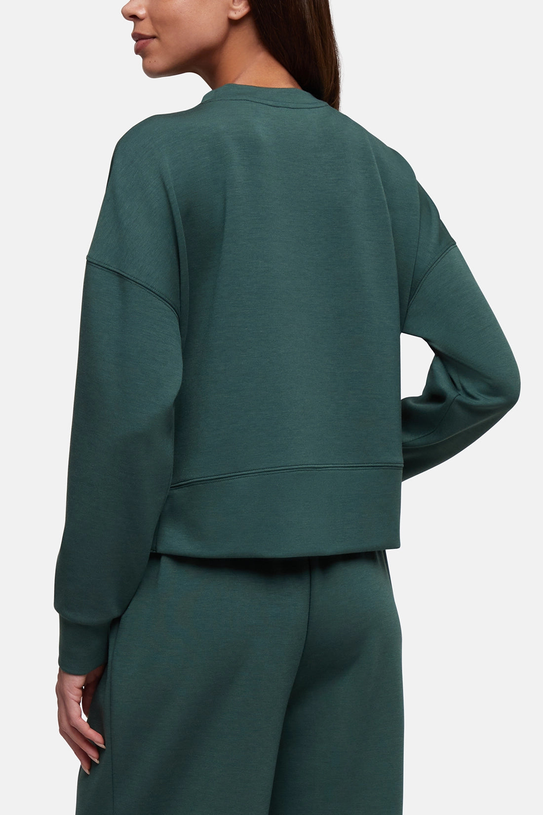 A woman shown from the back wearing a dark green cropped sweatshirt and matching sweatpants against a light gray studio background.