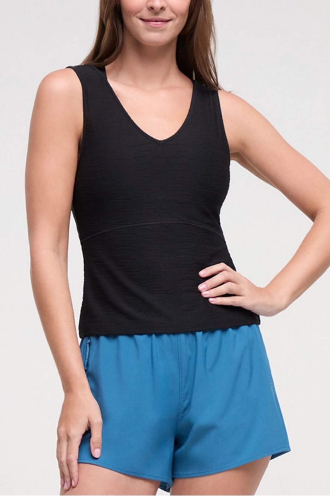 A woman wearing a black sleeveless V-neck athletic tank top and bright blue athletic shorts, shown from the shoulders to the upper thighs against a light gray studio background.