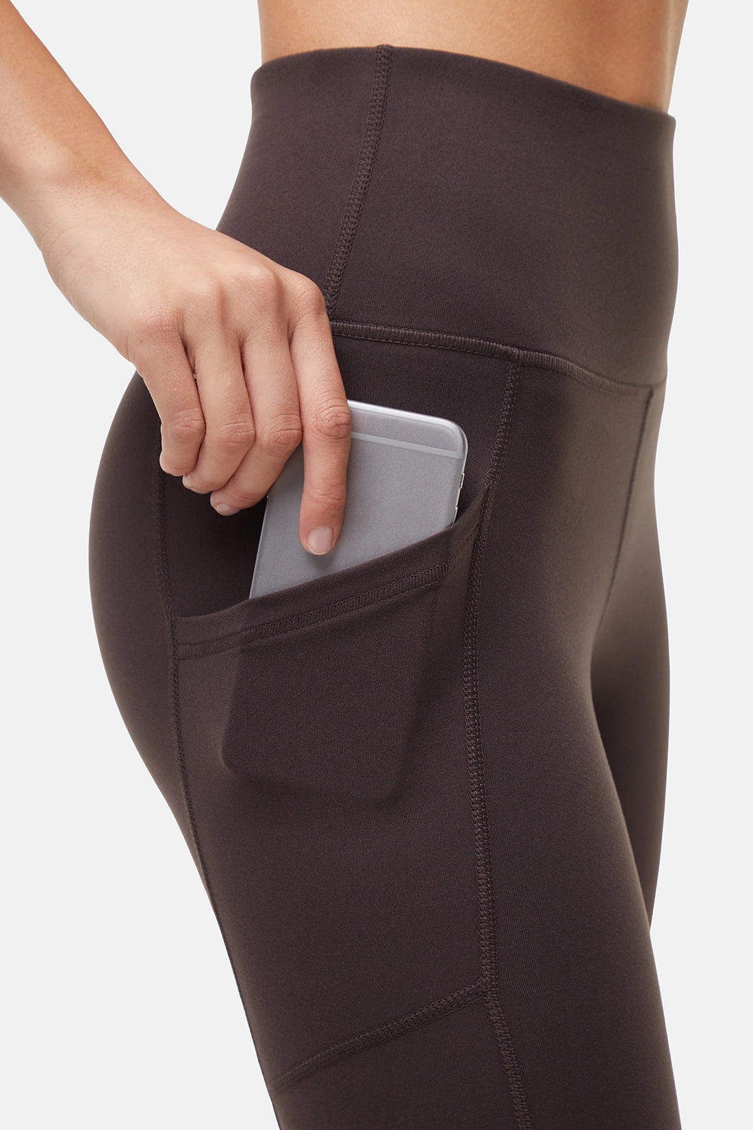 Woman wearing dark brown leggings with a side pocket holding a phone on a light gray background. 