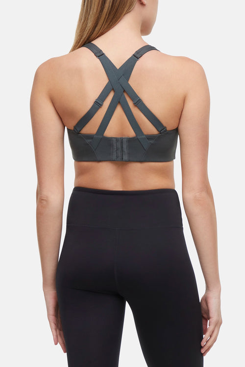 Back view of woman wearing a gray sports bra with criss-cross straps and black leggings on a white background. #color_urban-chic