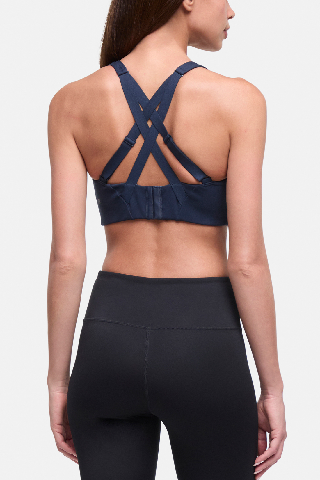 Rear view of a woman wearing a navy blue sports bra with crisscross back straps and black high-waisted leggings against a light gray studio background. 