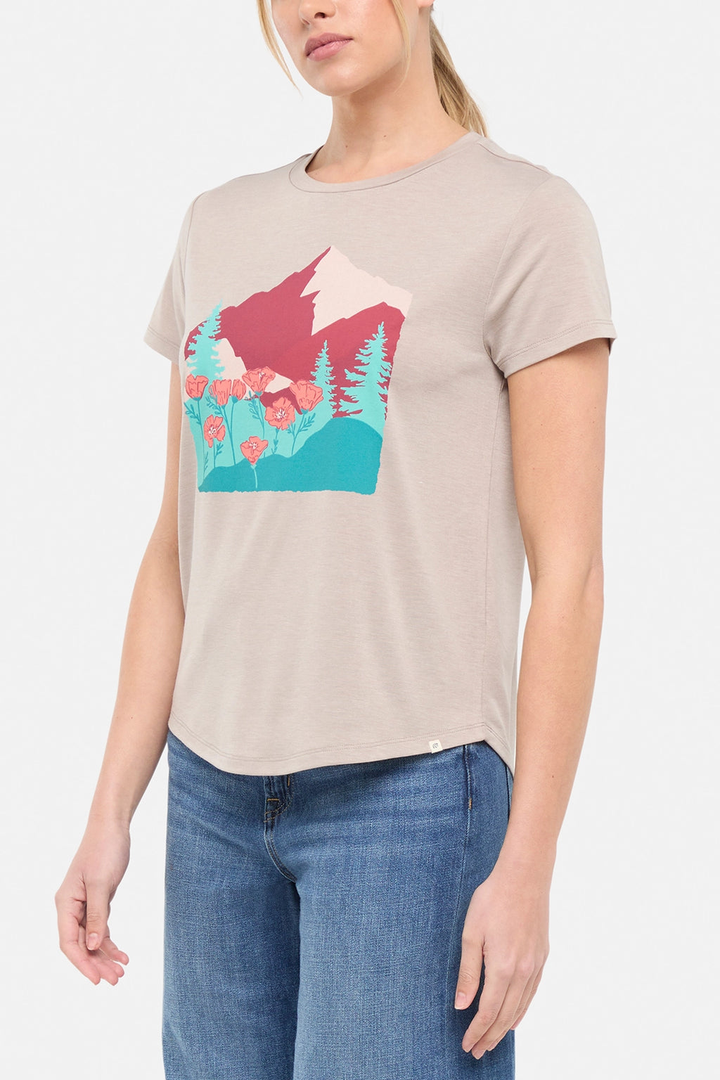 Side view of woman wearing a beige t-shirt with a graphic design of trees mountains, and poppy flowers, paired with blue jeans on white background. #color_heather-atmosphere-poppies-on-mountains