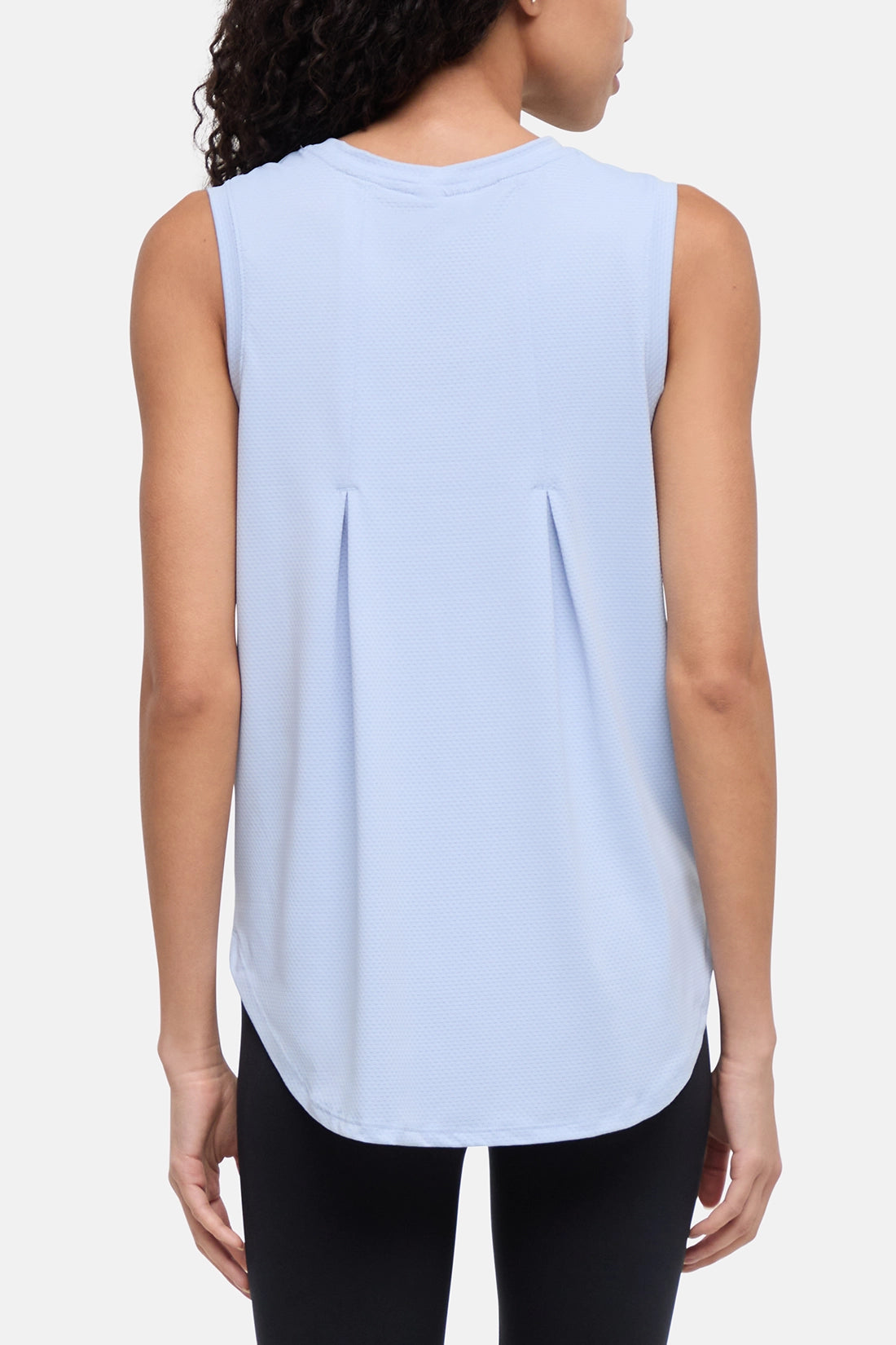 A woman shown from the back wearing a light blue sleeveless crop top with a curved hem and gathered seam detail, paired with black leggings, against a plain light gray studio background. 