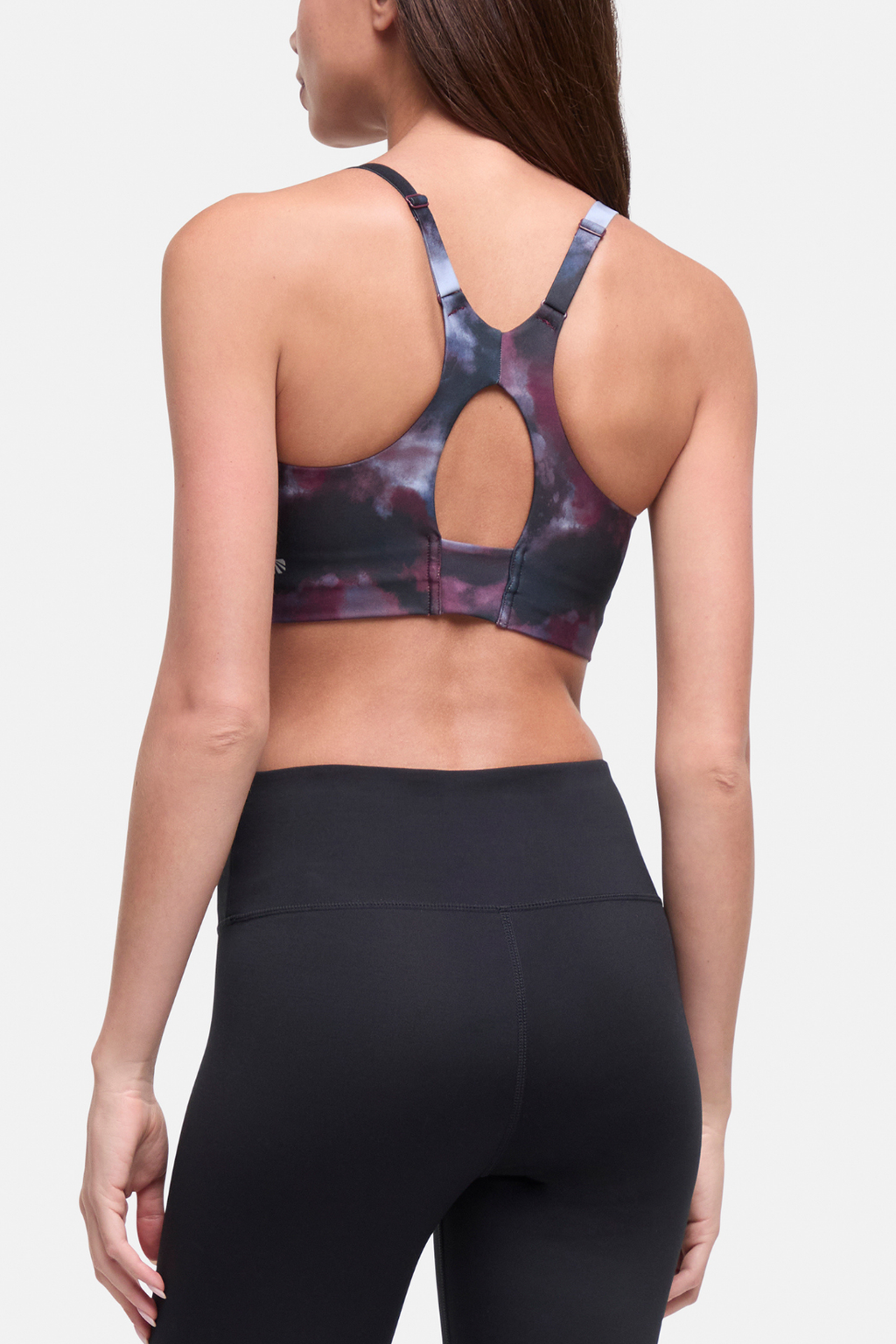 Rear view of a woman wearing a sports bra with a dark galaxy print and a keyhole cutout at the back, paired with black leggings, against a light gray studio background. #color_winetasting-clouded-cause
