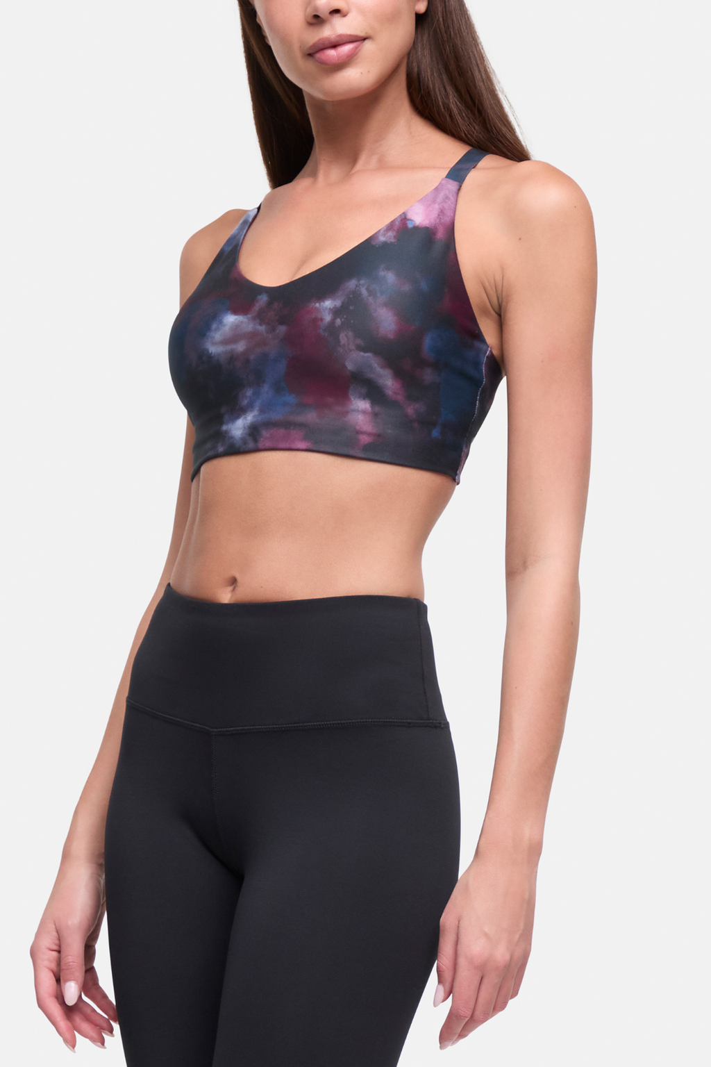 A woman modeling a sports bra with a dark navy, purple, and pink tie-dye pattern, paired with black high-waisted leggings, against a light gray studio background. #color_winetasting-clouded-cause