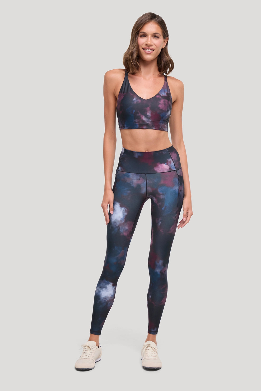 A woman modeling a matching activewear set featuring a dark galaxy print with purple, blue, and pink nebulae, consisting of a sports bra and high-waisted leggings, paired with beige sneakers, against a solid light gray background. #color_winetasting-clouded-cause