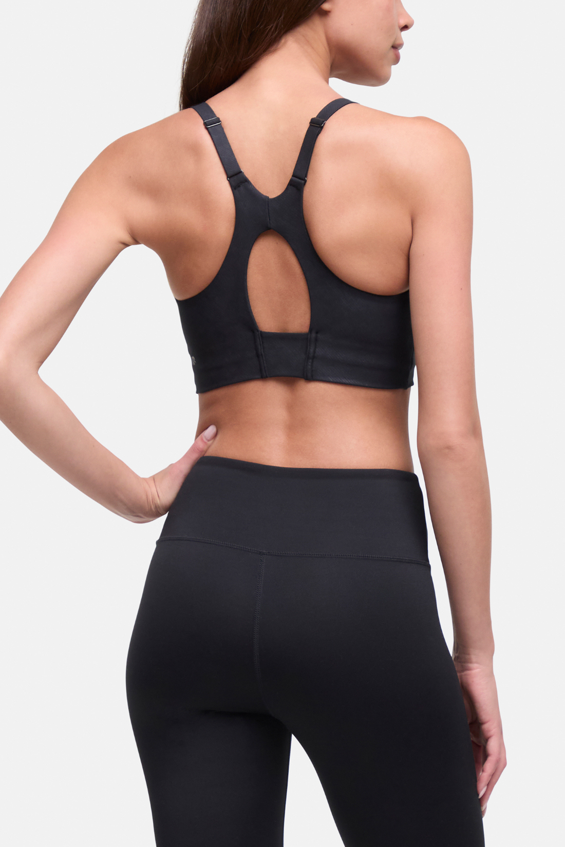 Rear view of a woman wearing a black sports bra with a cutout back and black high-waisted leggings against a light gray studio background. #color_black-linear-animal-camo
