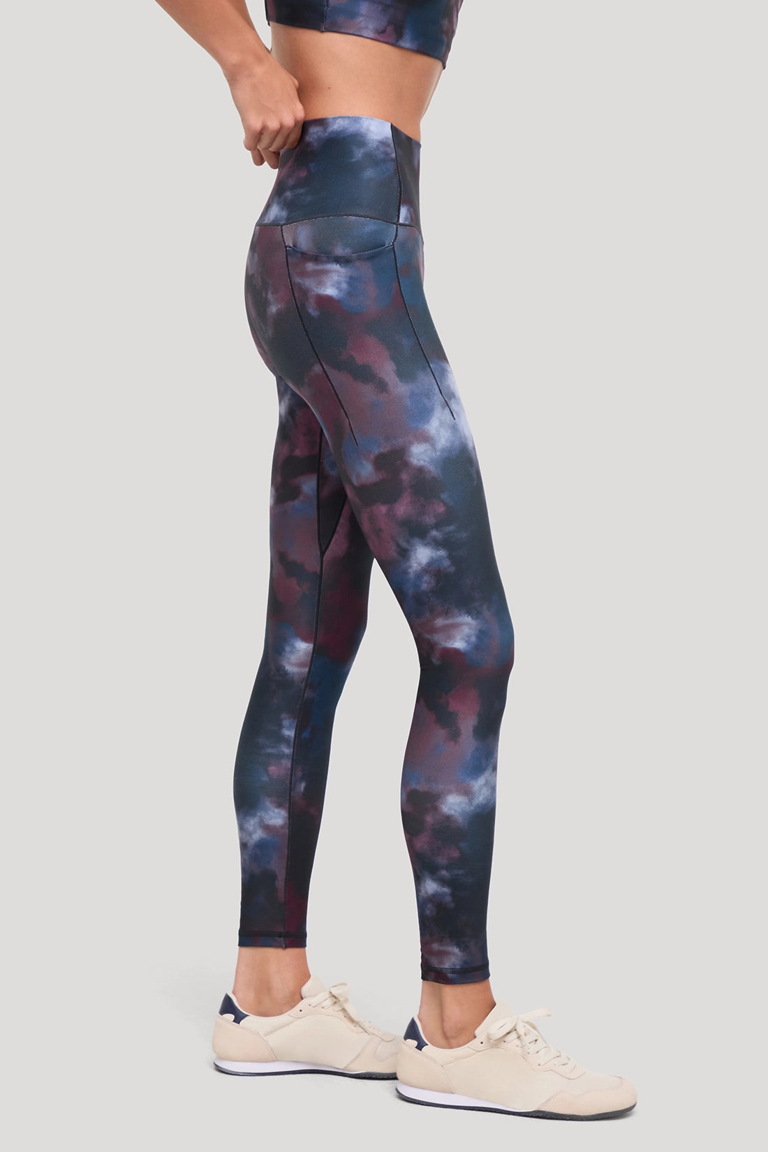 A model wearing high-waisted leggings with a dark blue, purple, and gray cloud-like tie-dye pattern, paired with beige sneakers, shown from the waist down against a solid light gray background. #color_winetasting-clouded-cause