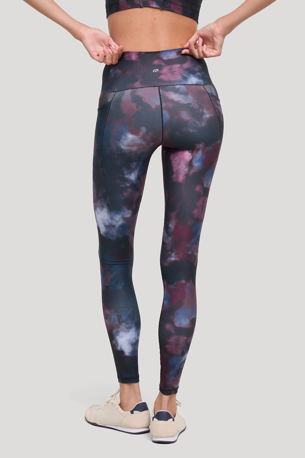 A model from the back wearing high-waisted leggings with a dark navy, purple, and blue tie-dye pattern, paired with beige sneakers, against a solid light gray background. #color_winetasting-clouded-cause