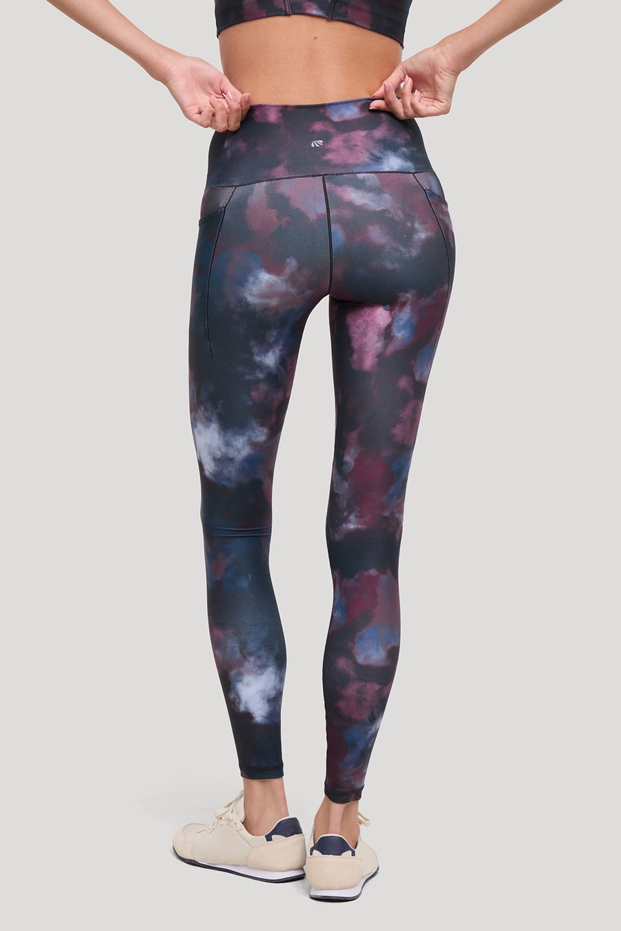 A model from the back wearing high-waisted leggings with a dark navy, purple, and blue tie-dye pattern, paired with beige sneakers, against a solid light gray background. #color_winetasting-clouded-cause