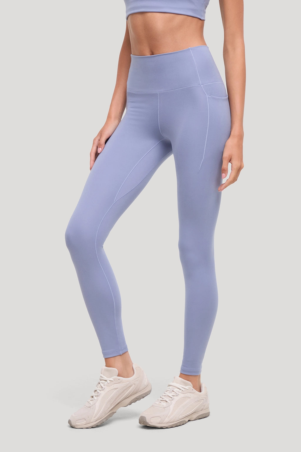 A model wearing high-waisted light periwinkle blue leggings and cream-colored athletic sneakers, shown from the waist down against a solid light gray background. #color_tempest