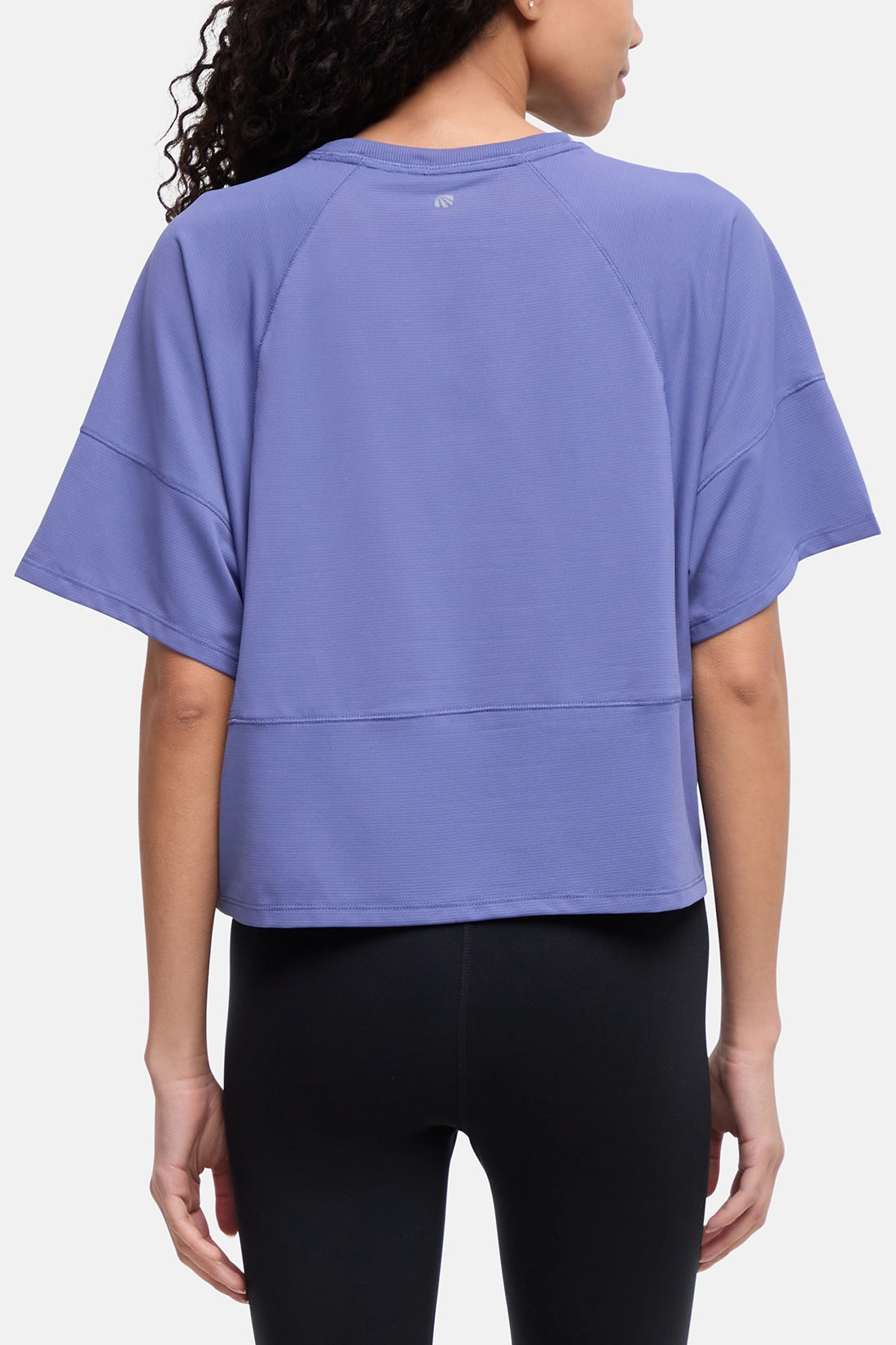 A woman shown from the back wearing a loose lavender-purple athletic top with raglan sleeves and black leggings, against a plain light gray studio background. 