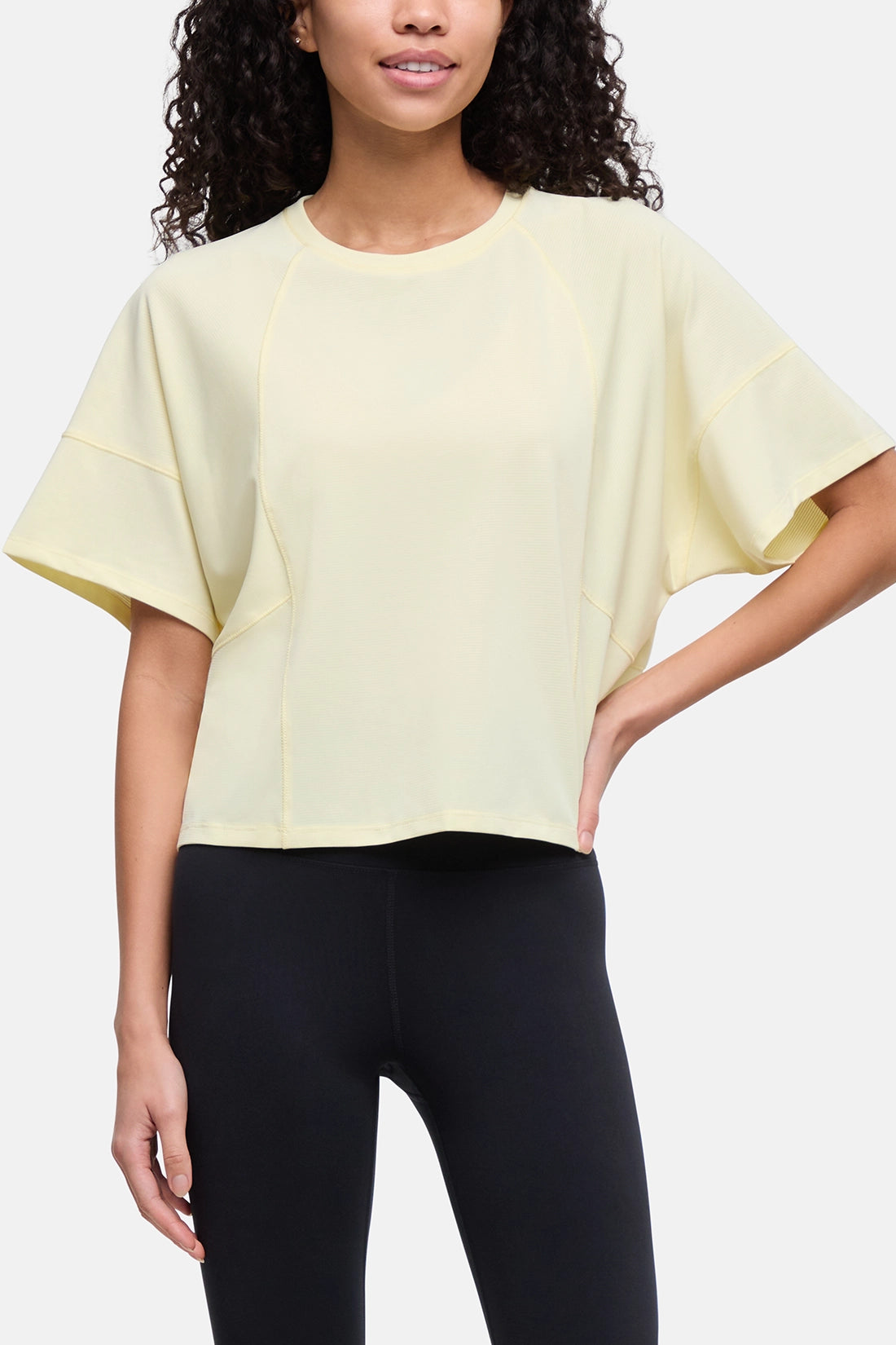 A woman wearing a pale yellow short-sleeve crop top with wide draped sleeves and black leggings, shown against a light gray studio background. 