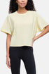 A woman wearing a pale yellow short-sleeve crop top with wide draped sleeves and black leggings, shown against a light gray studio background. 