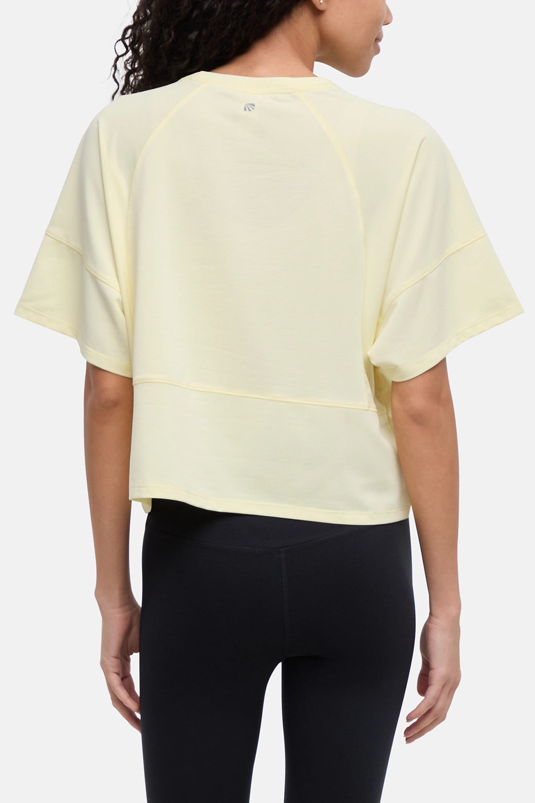 A woman shown from the back wearing a pale yellow short-sleeve cropped athletic top with raglan sleeves and black leggings against a light gray studio background. 
