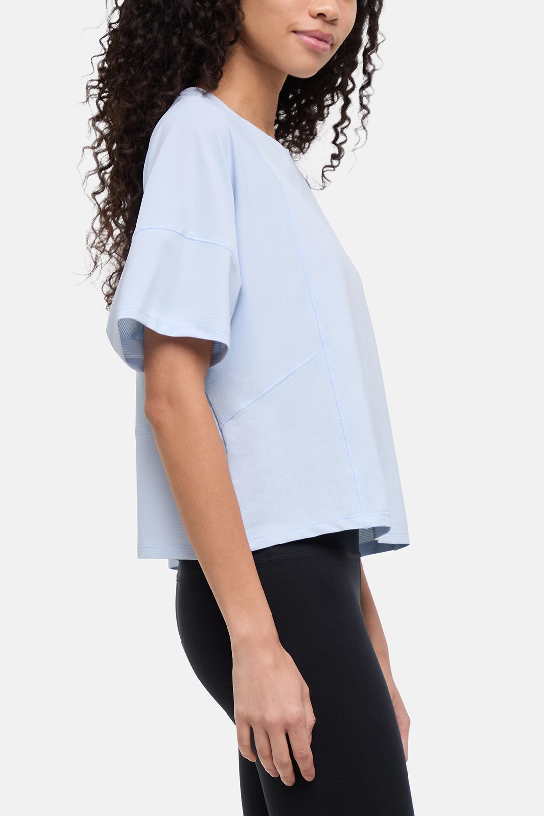 A woman wearing a light blue short-sleeve crop top with a relaxed fit and black leggings, shown from a side profile against a plain light gray background. 