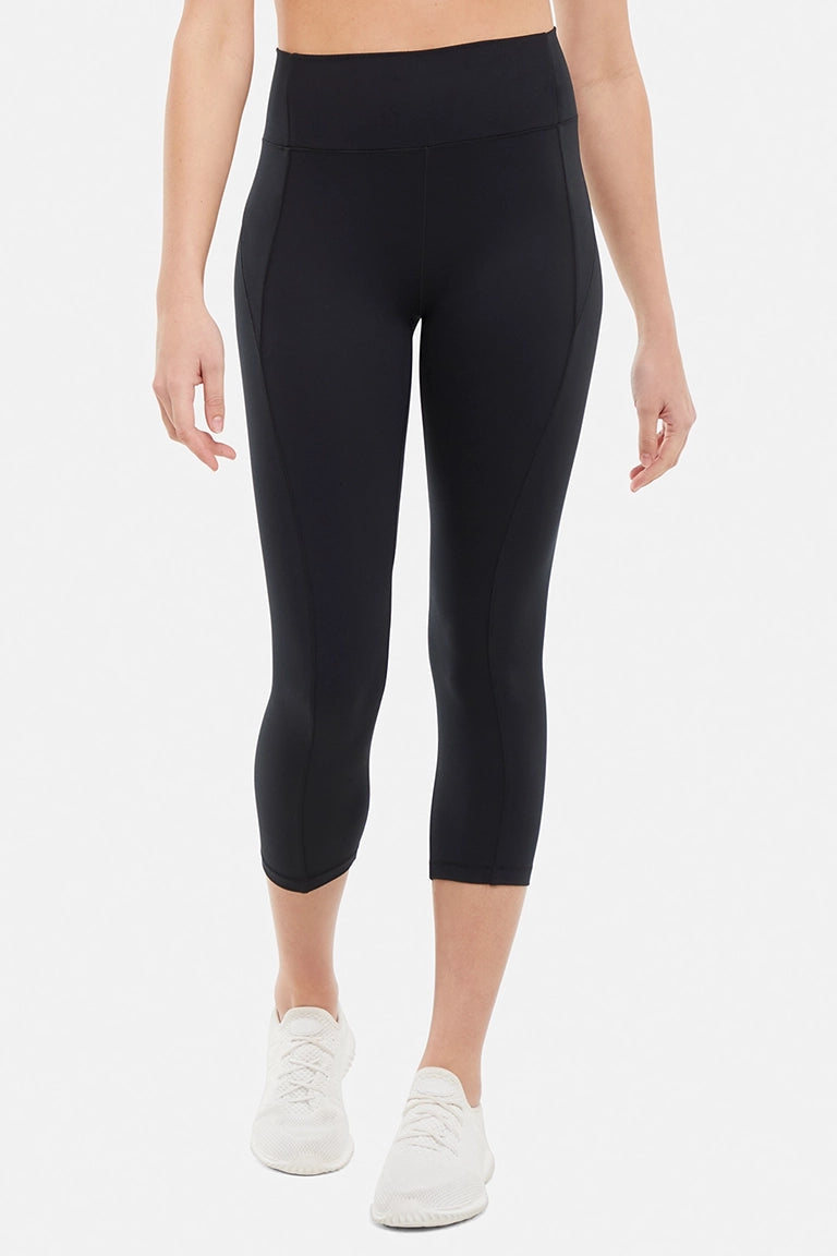 Front view of woman wearing black capris and white sneakers on a light gray background. #color_black