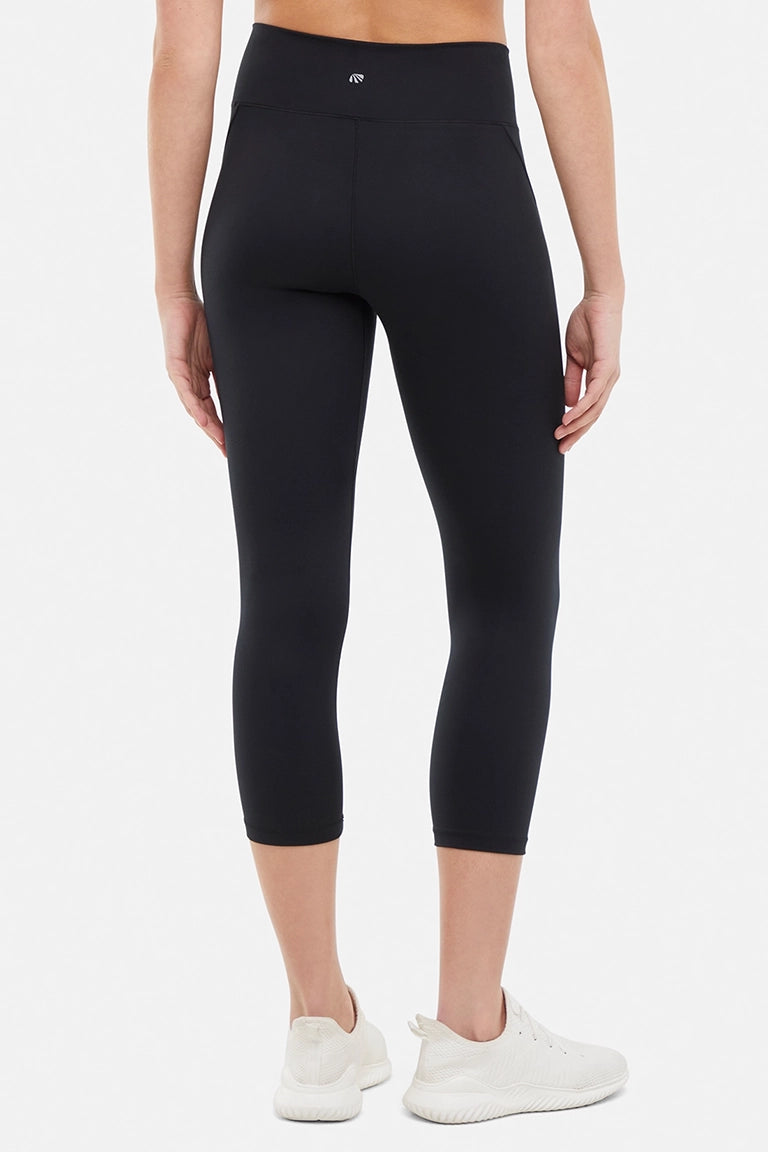 Back view of woman wearing black capris and white sneakers on a light gray background. #color_black