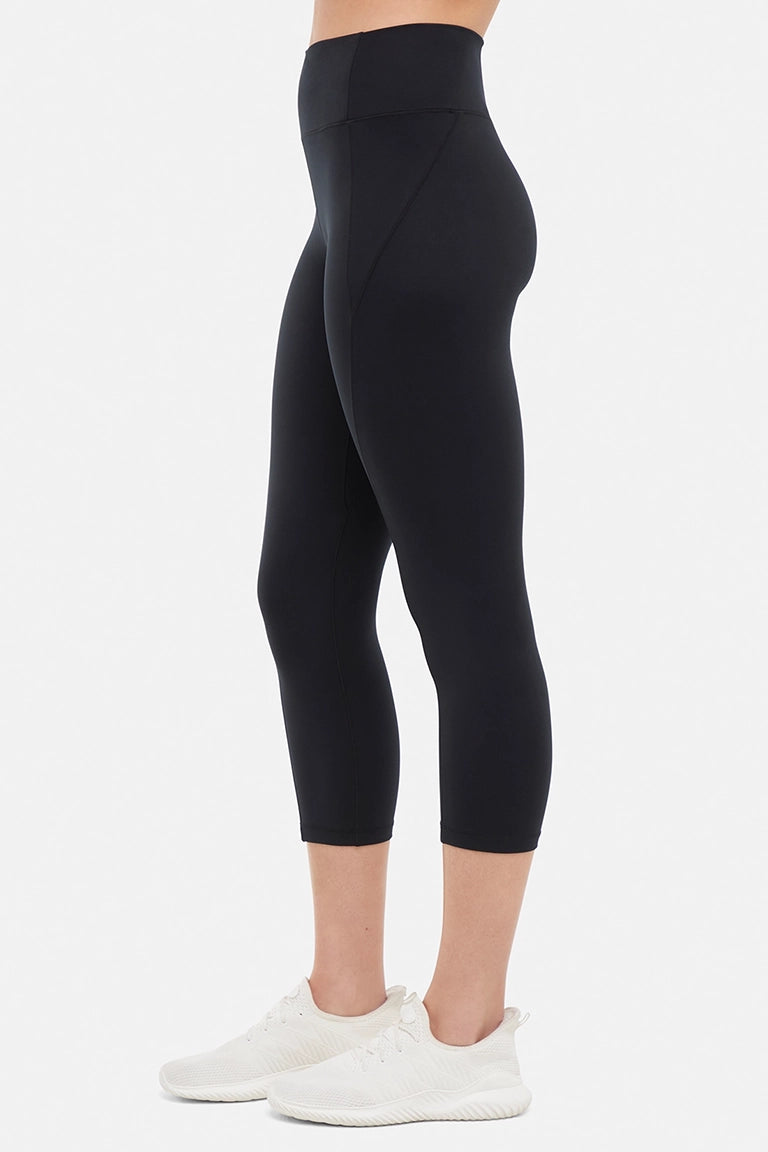 Side view of woman wearing black capris and white sneakers on a light gray background. #color_black