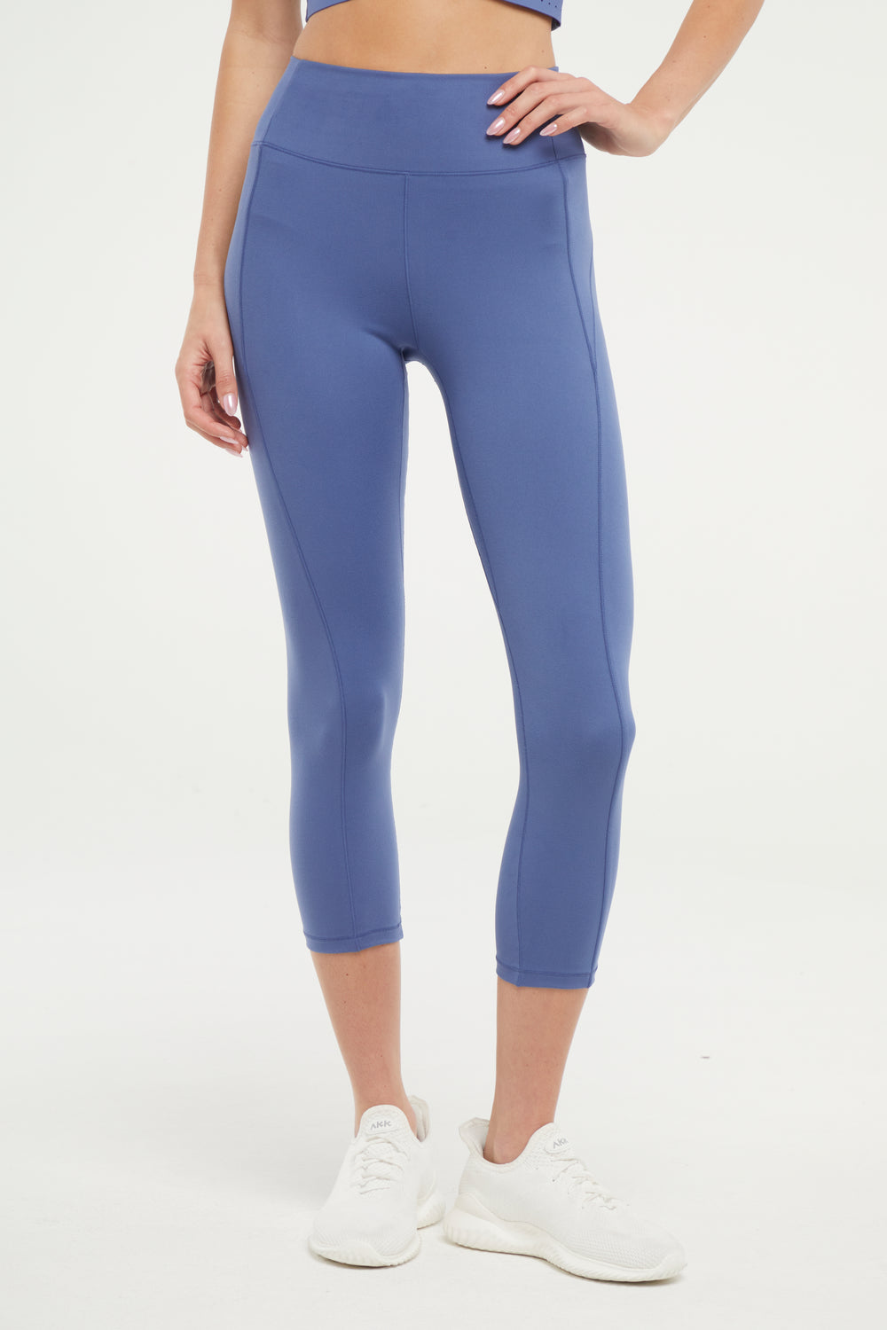 Front view of woman wearing blue capris and white sneakers on a light gray background..