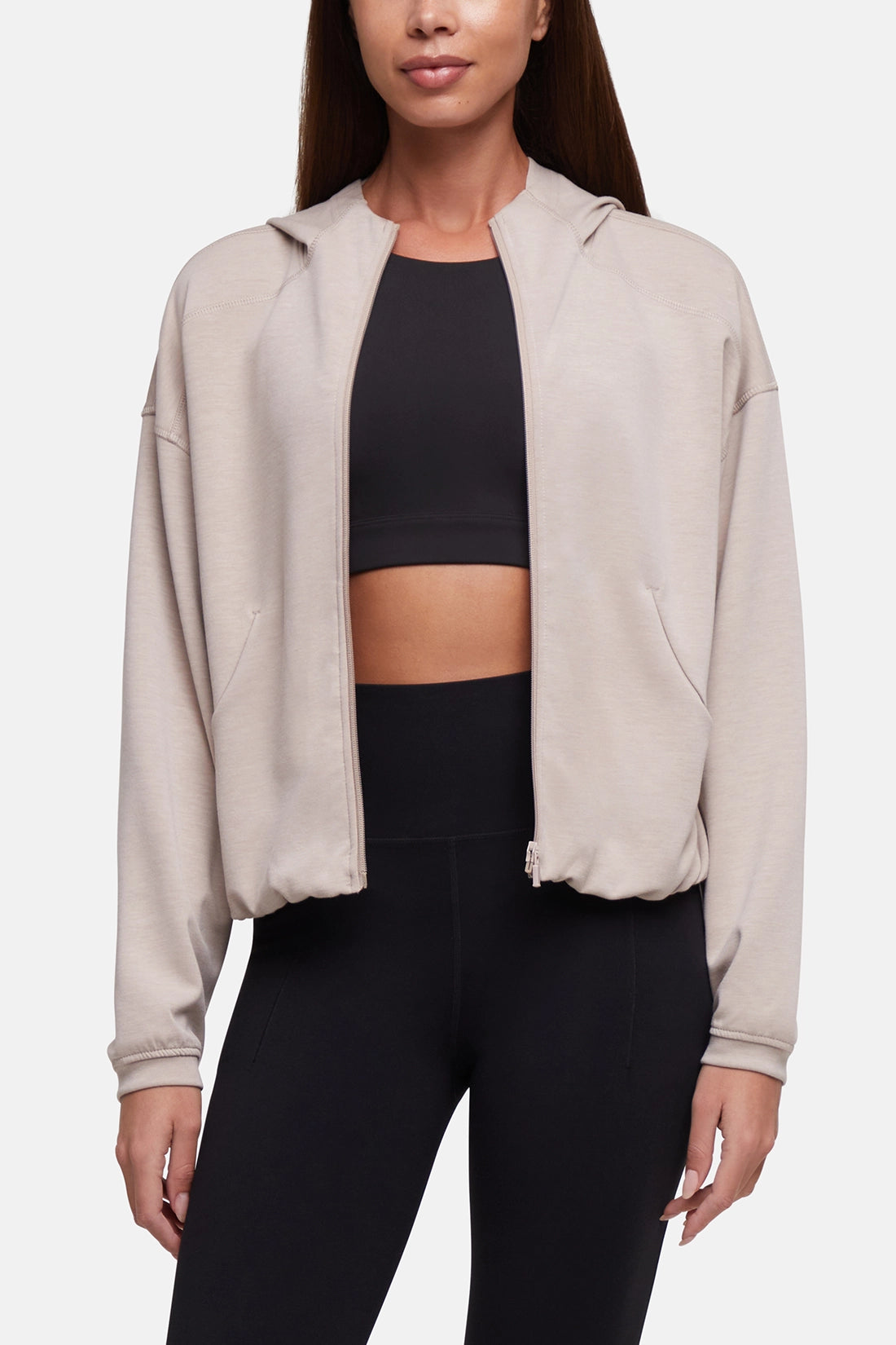 A woman wearing a light beige cropped zip-up hoodie with a hood and front pockets, layered over a black sports bra and black leggings, against a plain light gray studio background. #color_atmosphere