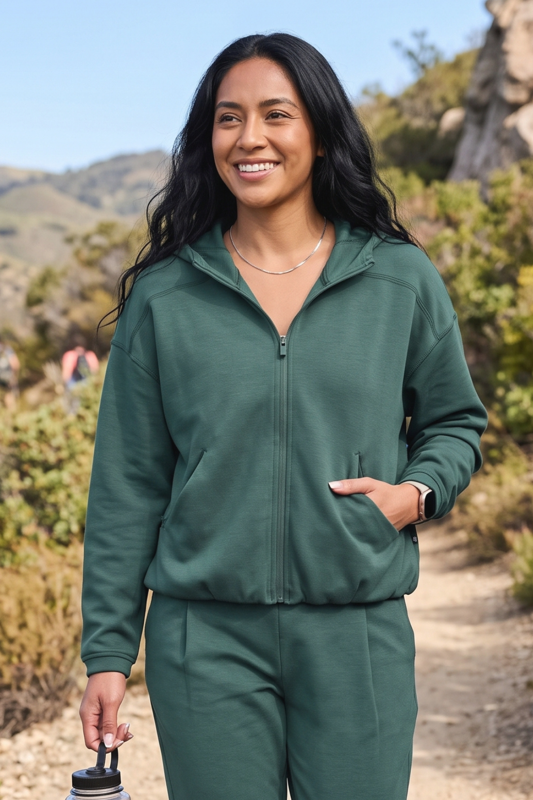 A woman wearing a matching dark green zip-up hoodie and sweatpants set stands outdoors on a dirt trail with hills and shrubs in the background. #color_garden-topiary