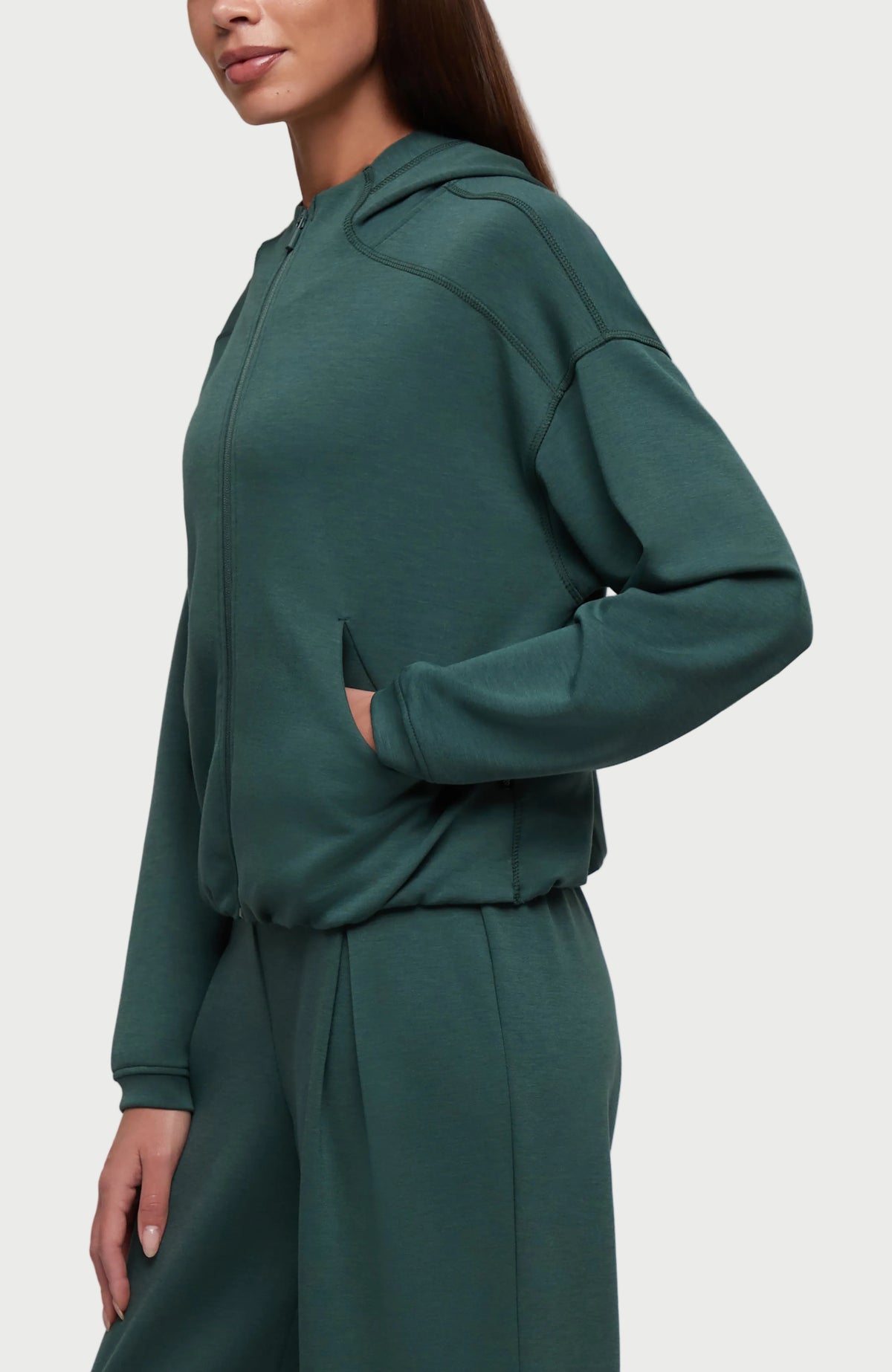 A woman wearing a dark teal green matching loungewear set, featuring a cropped zip-up hoodie with a kangaroo pocket and matching wide-leg sweatpants, shown against a plain white background. #color_garden-topiary