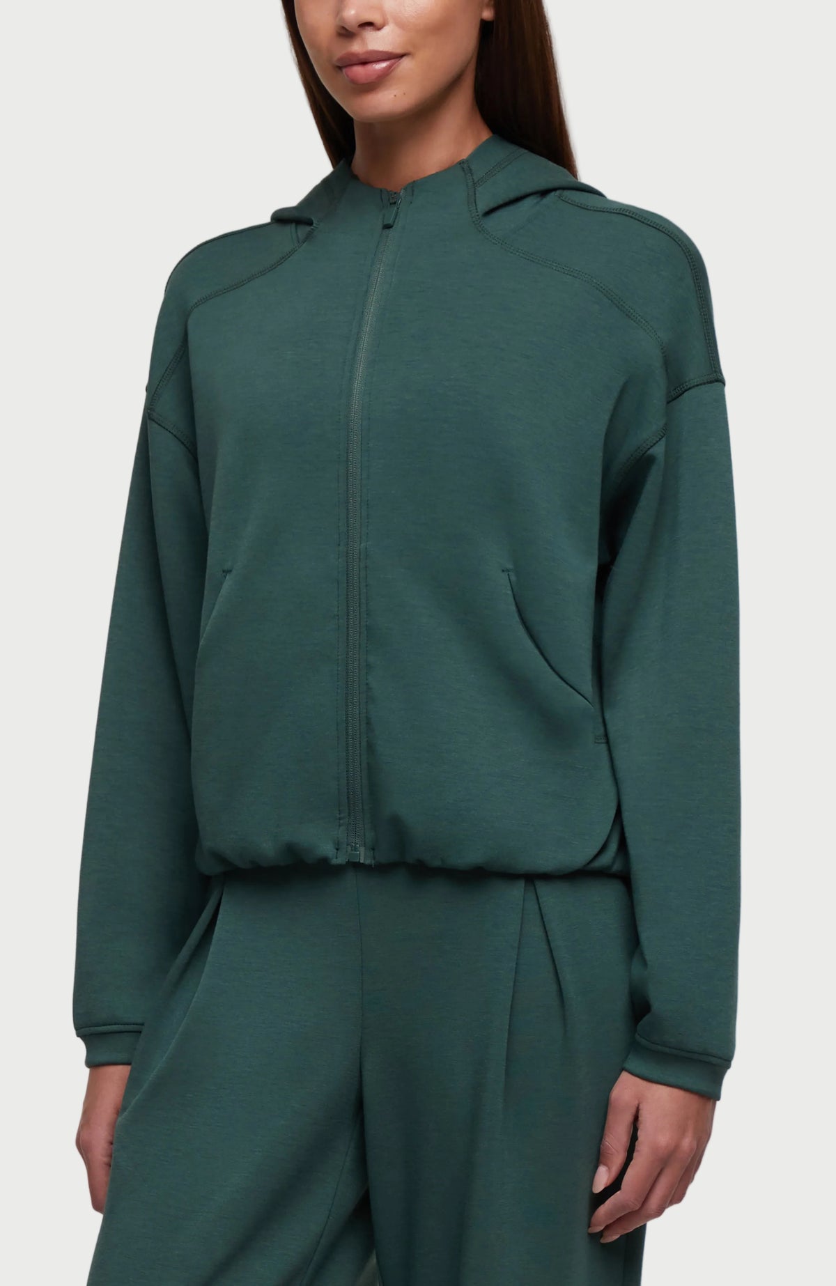 A woman wearing a dark teal green cropped zip-up hoodie with a matching pair of sweatpants, shown from the nose down against a plain white background. #color_garden-topiary