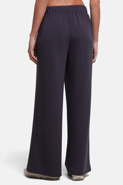 A person shown from the back wearing dark charcoal wide-leg pants with an elastic waistband, standing against a light gray studio background. 