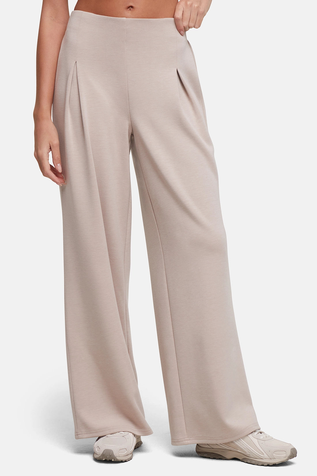 A model wearing high-waisted, wide-leg pleated trousers in a light taupe-beige color with front pleat detail, paired with light-colored sneakers, against a plain light gray studio background. 