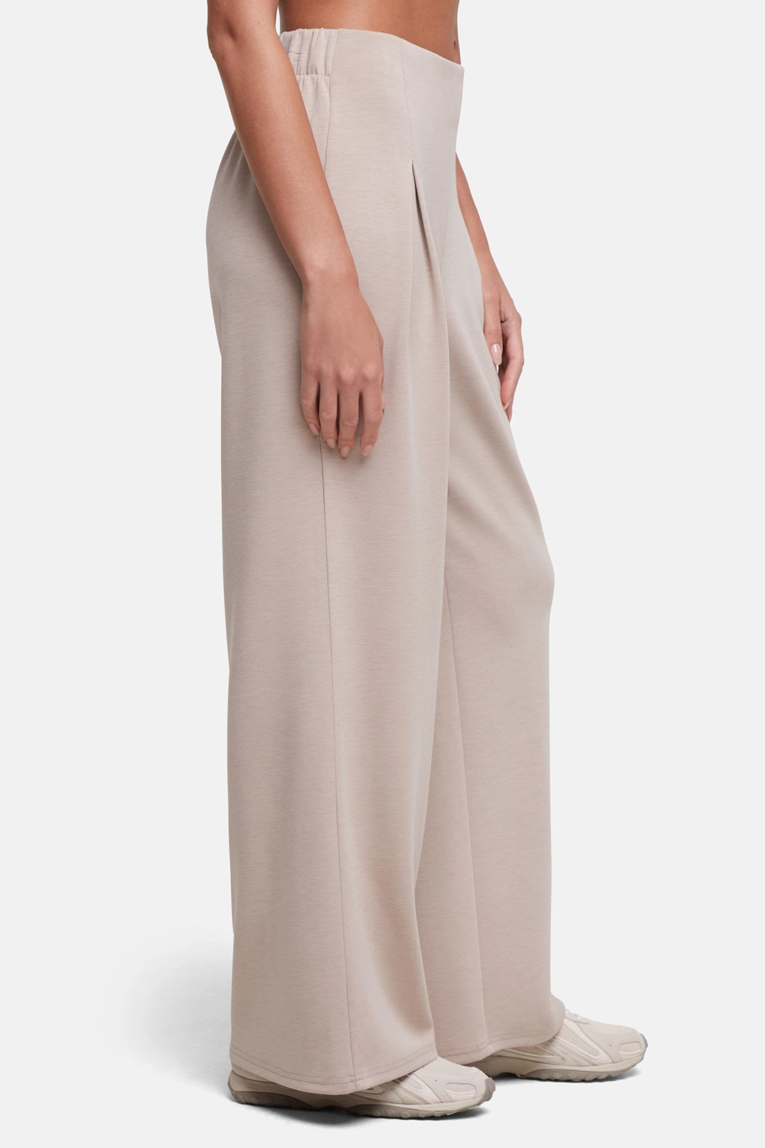 A model wearing high-waisted, wide-leg pleated trousers in a light beige or taupe color, paired with matching beige sneakers, shown from the waist down against a plain light gray studio background. 