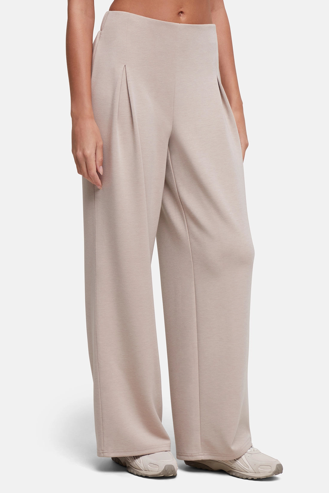 A model wearing a pair of wide-leg, high-waisted trousers in a light taupe-beige color, paired with neutral sneakers, against a clean light gray studio background. 