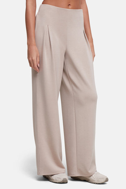 A model wearing a pair of wide-leg, high-waisted trousers in a light taupe-beige color, paired with neutral sneakers, against a clean light gray studio background. 