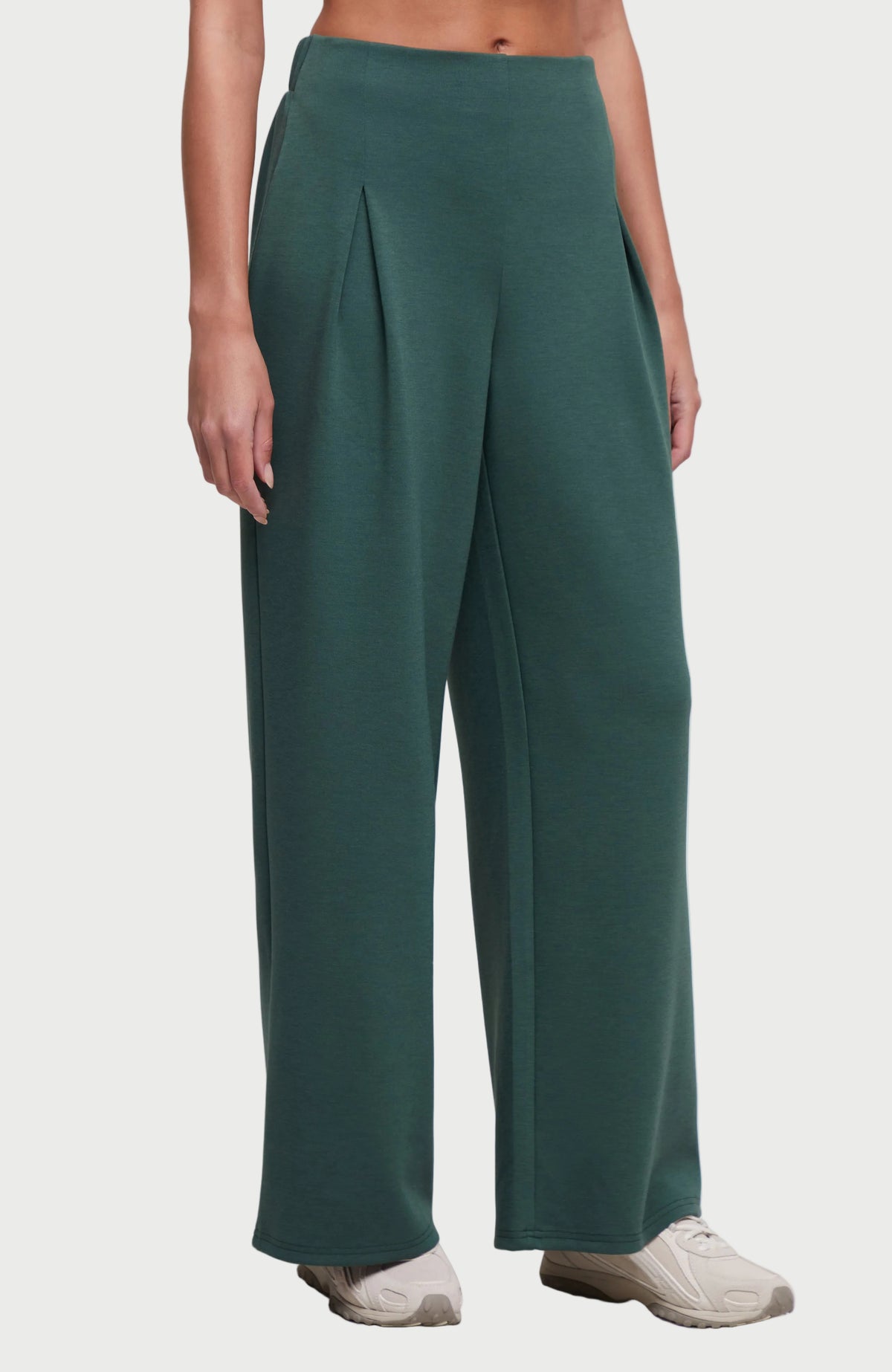 A model wearing a pair of wide-leg, high-waisted pants in a deep teal green color, paired with light gray sneakers, shown from the waist down on a grey studio background. 