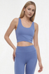 A woman modeling a matching athletic set consisting of a scoop-neck crop top and high-waisted leggings in a dusty periwinkle blue color, shot against a plain white studio background. 