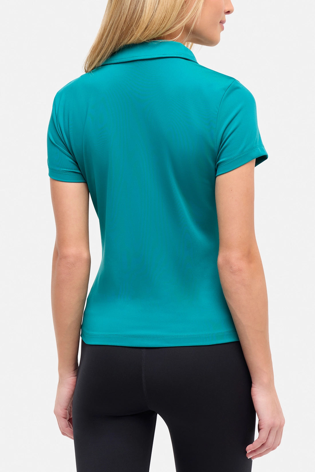 Back view of woman wearing a teal polo shirt and black pants on a white background. 