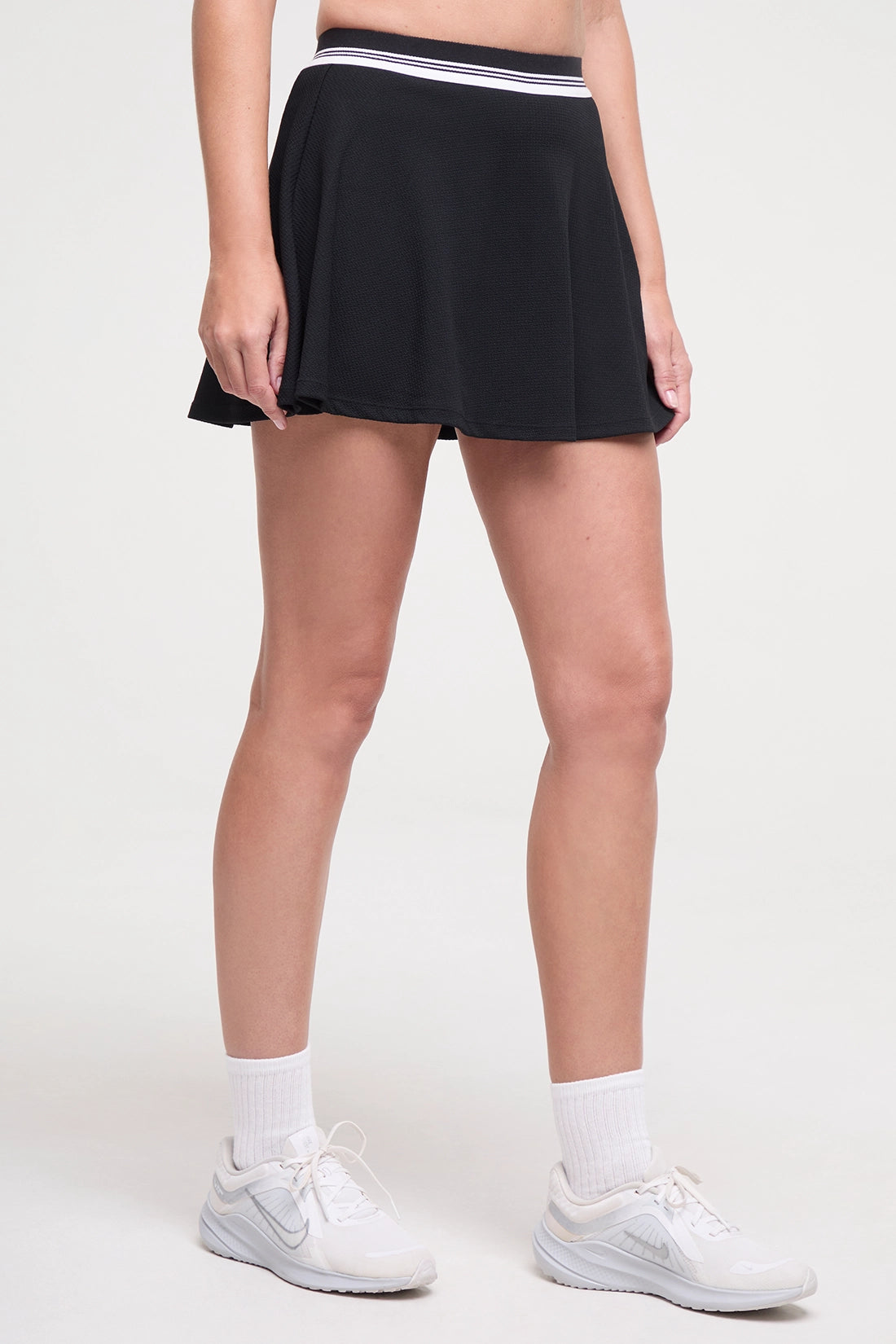 Side view of black tennis skirt with white stripe waistband worn by a woman on a white background. 