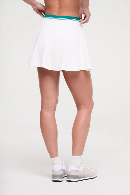 Back view of white tennis skirt with teal stripe waistband and inner short pocket worn by a woman on a white background. 